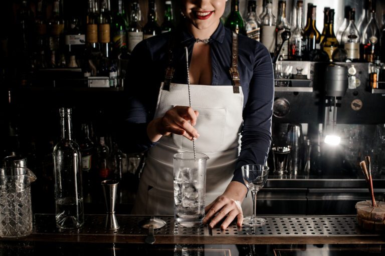 Smiling bartender woman stirring cocktail with ice in a glass on the bar counter