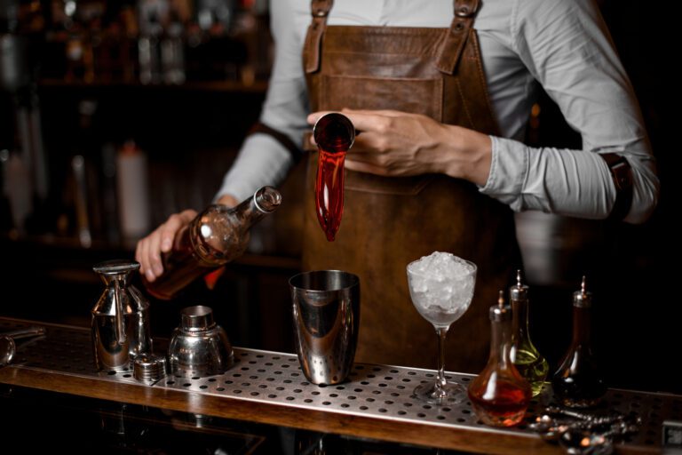 Male bartender pouring a red alcoholic drink from the steel jigger to the cocktail shaker on the bar counter in the dark blurred background