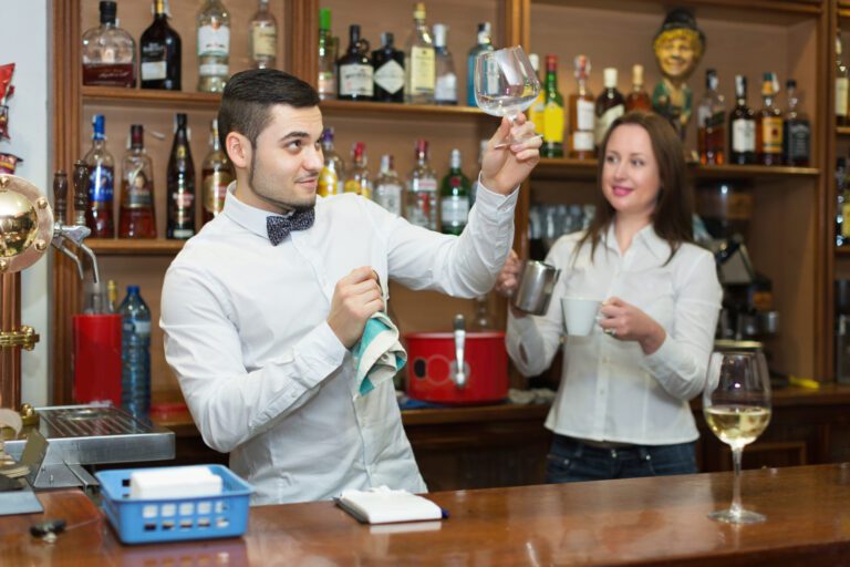 Positive waitress and smiling barmen working in modern bar. Focus on man inspecting a glass for cleanliness.